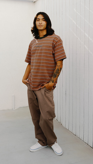 A young man posing outside wearing an Obsidian Fern-Light Bronze Cameron Stripe Knit Shirt and white Authentic shoes