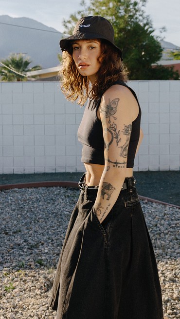 Une jeune femme pose à l’extérieur. Elle porte une jupe Authentic Chino en jean noir, un chapeau de pêcheur Dropped V noir et une camisole New Salton noire.