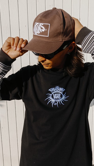 A young man wearing a black Torched T-Shirt T-Shirt and a vintage cocoa brown Vans Formula Snapback Hat.