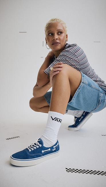 A young woman squatting in a photography studio wearing navy and marshmallow white Authentic Boat shoes and a blue and gray Cameron Stripe Knit Shirt.