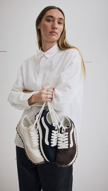 A young woman holding all three marshmallow white, black, and demitasse brown colorways of the Premium Style 152 by their laces in a photography studio.