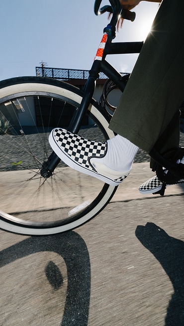 A knee-down shot of someone biking in the sunlight wearing black and white Checkerboard Classic Slip-On shoes.