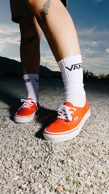 Close-up of someone wearing orange Authentic shoes with white Vans crew socks on gravel at dusk.