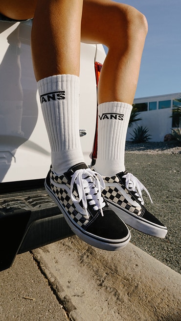 A knee-down shot of someone sitting on the gate of a truck wearing black and white Old Skool Checkerboard shoes and white Vans crew socks.