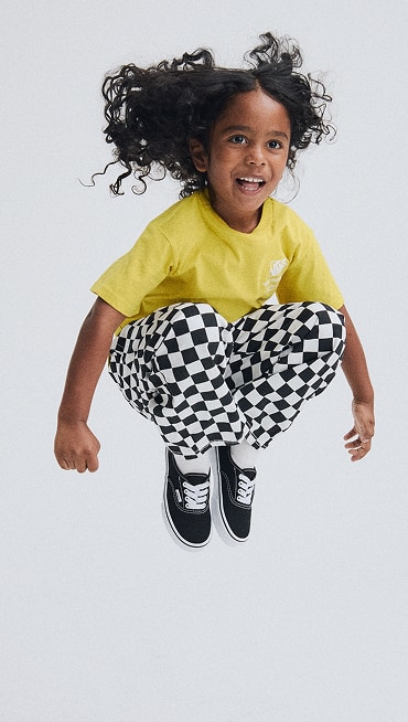 A young girl jumping and wearing a yellow Stacked Hi T-shirt, black and white Lili Elastic Waist Checkerboard Pants, and black and white Authentic.