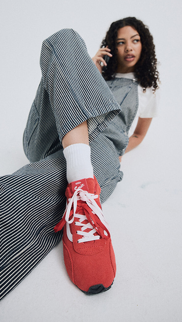 A young woman leans back on the floor of a photography studio wearing racing red Super Lowpro and Hickory Stripe Blue Chore Stripe Overalls.