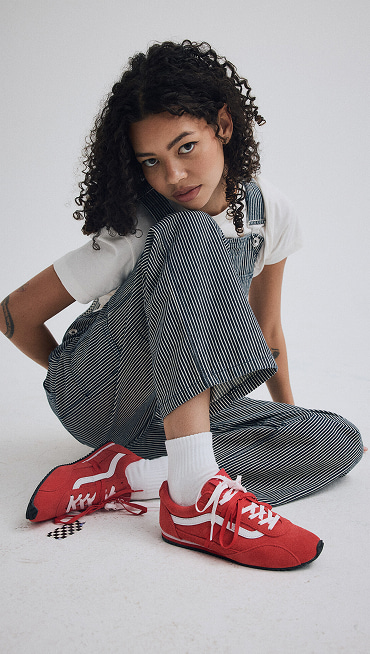 A young woman sitting on the floor of a photography studio wearing racing red Super Lowpro and Hickory Stripe Blue Chore Stripe Overalls.