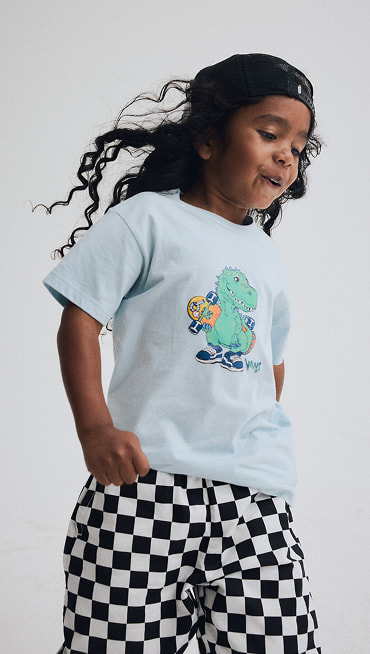 A young boy wearing black and white Checkerboard Little Kids Liam Elastic Waist Pants and a dawn mist blue Little Kids Prehistoric Skater T-Shirt.