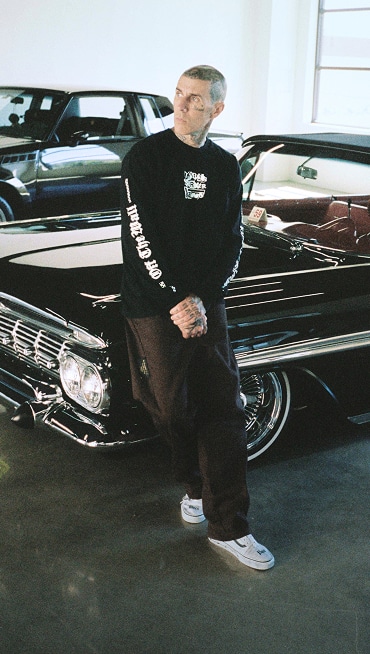Blink-182 drummer leans up against a black vintage car in a garage wearing the white Vans X Travis Barker Old Skool.