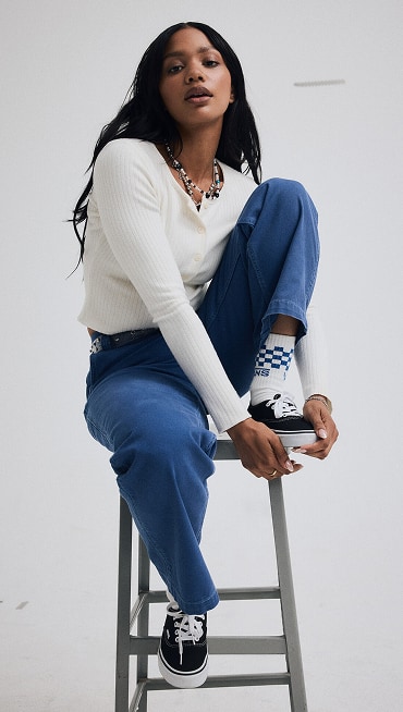 A young woman sits on a stool in a photo studio wearing a marshmallow white Long Sleeve Button Up Shirt and black Authentic shoes.