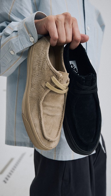 Close-up on someone holding the light brown Premium SF Moc Low Shoe and a black Premium SF Moc Low Shoe.
