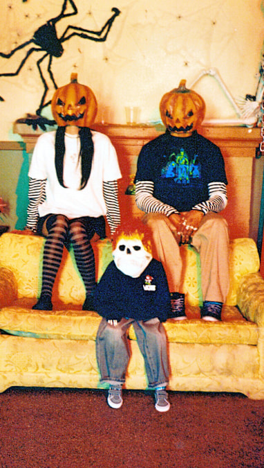 Two people and a skeleton in masks wear Vans apparel and footwear, sitting on a yellow couch.
