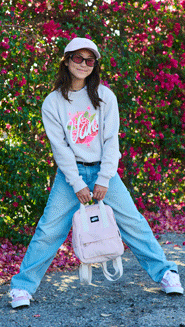 A young girl poses with her sepia rose pink Old Skool Classic Backpack and Vans gray pullover in front of a rose bush.