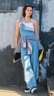 A woman leans up against a wall wearing denim blue Sirelle Overalls, a white tanktop, and black and white Platform Old Skool.