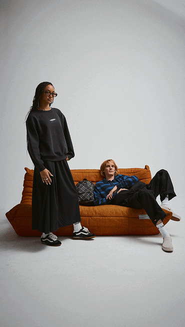 A young man and woman sit on an orange couch wearing beige Slip-On and black Super Lowpro in a photography studio.