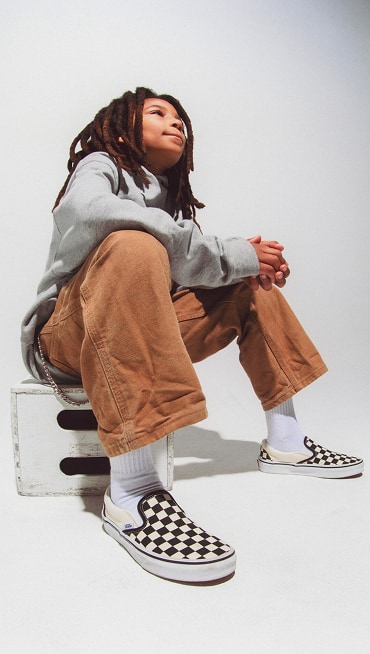 A kid sitting down wearing Classic Slip-On Checkerboard Shoes.