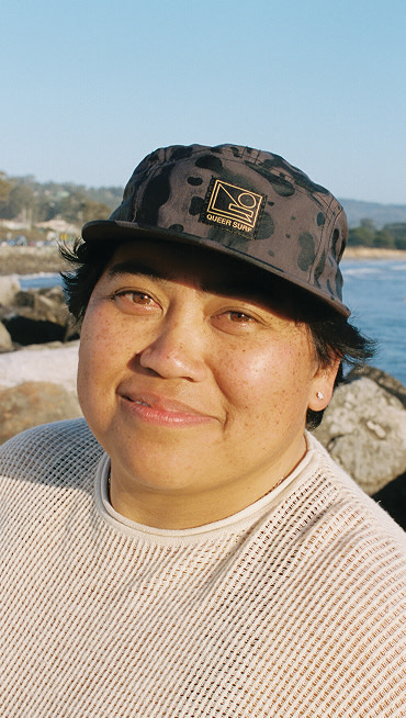 A person smiles into the camera with a Vans and Queer Surf bucket hat with a beach in the background.