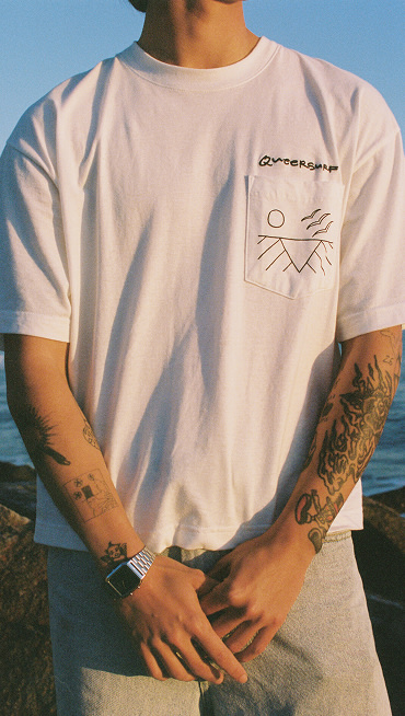 A close-up of someone wearing a white t-shirt with the phrase "Queer Surf" over the pocket.