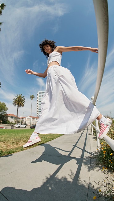 A woman jumps from a rail wearing a white crop top, a white dress, and Honeysuckle Pink Checkerboard Classic Slip-On near a beach.