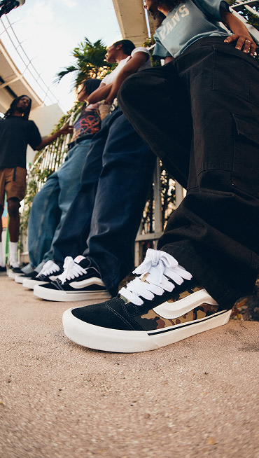 A close-up of a pair of camo Knu Skool on a man wearing black jeans with a group of young people in the background under a tram line.