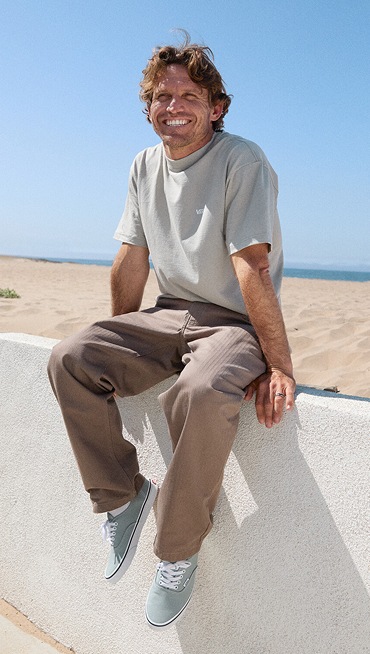 A man sits on a wall wearing Pure Grey Authentic shoes, Skate Loose Pleated Pants, and Vans t-shirt.