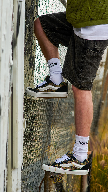 A waist-down shot of a young man climbing a cyclone fence wearing black Drill Chore Loose Denim 22-inch Shorts and camo Knu Skool.