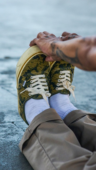 Close-up of a person’s feet as they stretch by pulling at the toes of their Vans Premium Authentic shoes in Antique Gold and Green color.