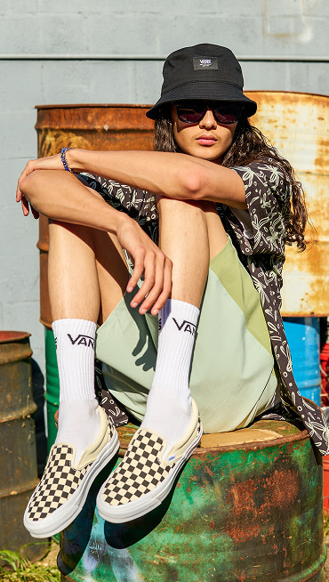 A guy poses on a graffied oil drum, wearing iconic checkerboard Classic Slip-On shoes styled with Vans Bucket Hat, sunglasses, and groovy button up.