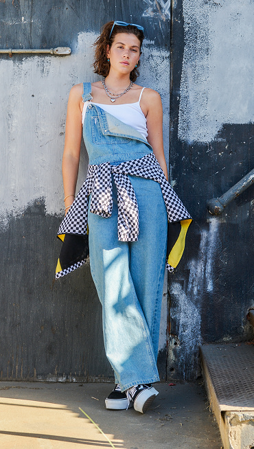 A woman leans against an exterior wall, wearing denim overalls and classic Vans Old Skool shoes, with a checkered windbreaker tied around her waist.