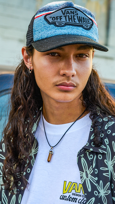 A man with long hair looks at the camera wearing a black and mint Hawaiian shirt and a Vans Off the Wall trucker hat.