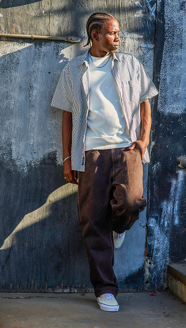 A man leans on a wall wearing a blue and white Lawson Stripe Button Up Shirt, brown jeans, and white and blue Premium Authentic.
