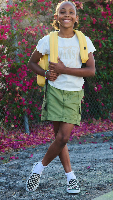 A young girl poses with her banana cream yellow Kids Old Skool Grom Backpack and Loden Green Kids Sidewalk Cargo Skirtin front of a rose bush.