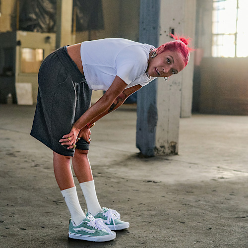 A woman with vibrant red-dyed hair rests with her hands on her knees in a warehouse wearing white Vans crew socks and mint green Knu Skool.
