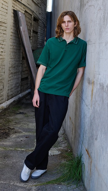 A man with long red hair poses in an alley wearing a rain Forest Green Skate Mesh Polo Shirt, dark pants, and gray Curren shoes.n shot of a man skating in a crosswalk wearing blue jeans and Skate Old Skool shoes with the city in the background.