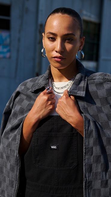 A woman poses on the street wearing black Sirelle Overalls and a gray and black Checkerbord jacket.