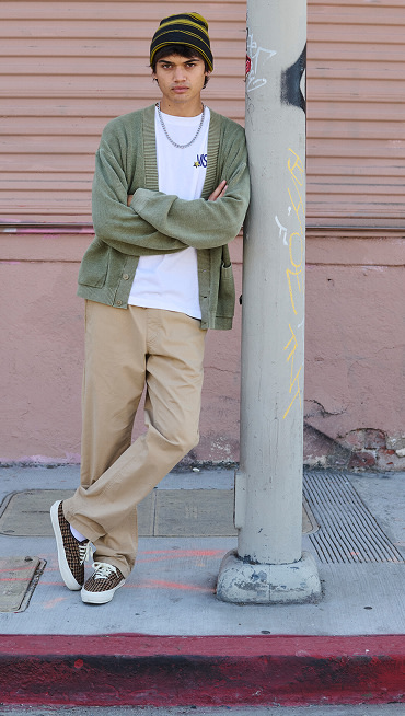 A young man scowls at the camera wearing engineered knit Authentic shoes and a moss green cardigan as he leans against a metal pole.