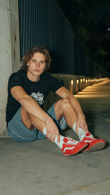 A man sits on the concrete and looks into the camera wearing a black t-shirt, denim blue jeans, and Racing Red Super Lowpro.