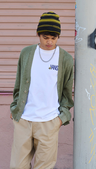 A young man looks at his feet wearing a white Vans t-shirt and a moss green cardigan as he leans against a metal pole.