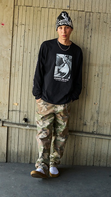A young man poses in a warehouse wearing camo pants, the Hovley beanie, and white Premium Authentic.