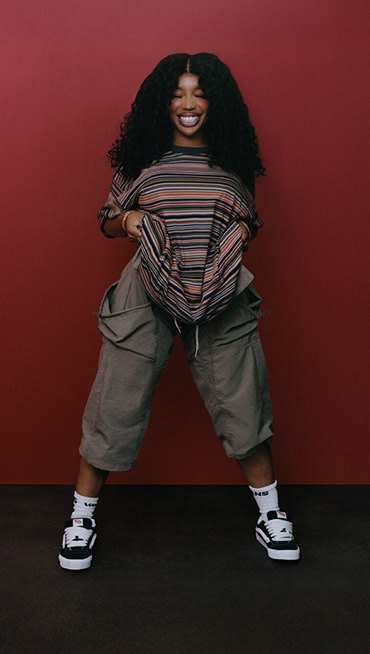 Singer SZA poses in a red room and smiles with her eyes closed wearing a striped t-shirt, and black and white Knu Skool shoes.