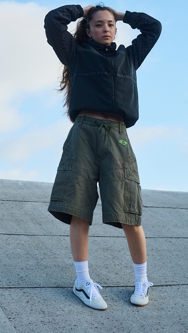 A young woman poses on a roof wearing a black hoodie, green cargo shorts, and marshmallow white Super Lowpro.
