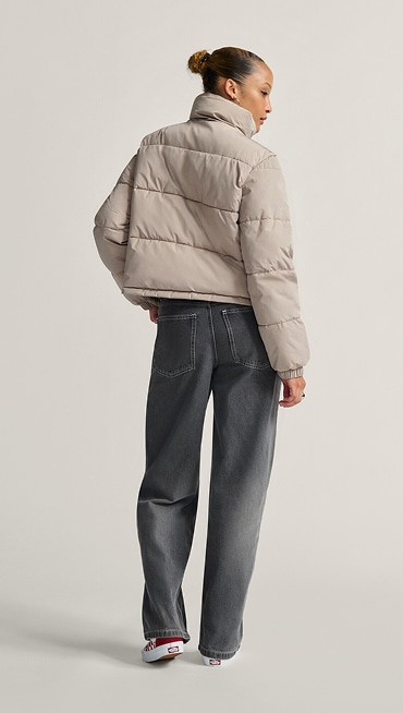 A young woman poses in a photography studio wearing a light brown MTE Aubrey Primaloft Puffer and gray Sirelle 5 Pocket Puddle Pants.