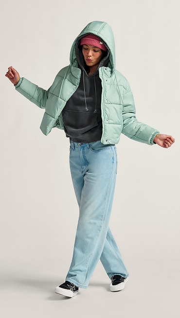 A young woman dances in a photography studio wearing a light blue MTE Hillgate Puffer, a Core Basic Cuff Beanie, and Cinched Check 5 Baggy blue jeans.