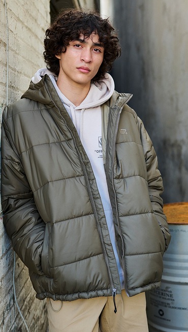A man poses in a warehouse and looks into the camera wearing a taupe mist Stockpile Pullover Hoodie and brown MTE Hillgate Puffer.