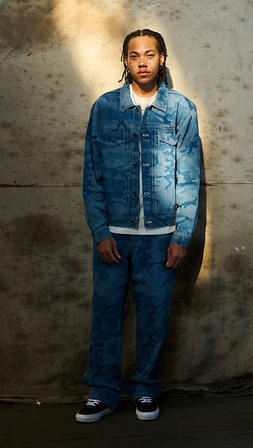 A man poses in a warehouse wearing a stonewash blue Camo Denim Trucker Jacket and Check 5 Loose Camo Printed Denim Pants.