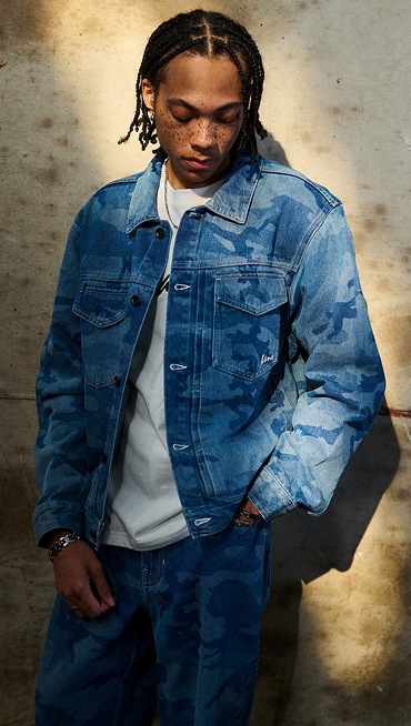 A man poses in a warehouse wearing a stonewash blue Camo Denim Trucker Jacket and Check 5 Loose Camo Printed Denim Pants.