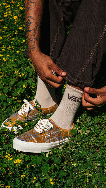 A knee-down shot of someone lacing up their Premium Authentic Harris Tweed shoes paired with gray Vans crew socks in a park.