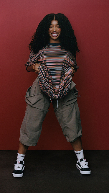 Singer SZA poses in a red room and smiles with her eyes closed wearing a striped t-shirt, and black and white Knu Skool shoes.