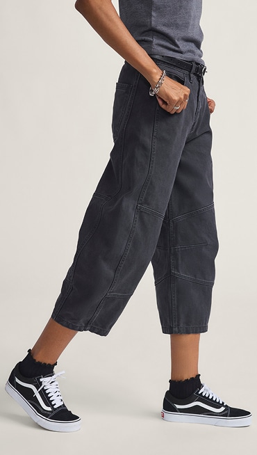 A waist-down shot of a young woman wearing black Emily DIY Pants with black and white Old Skool in a photography studio.
