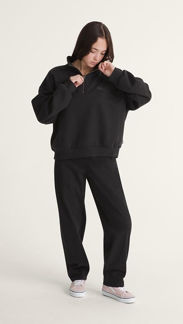 A young woman poses in a photography studio wearing a black Script Logo Sweatshirt, black pants, and pink Old Skool.